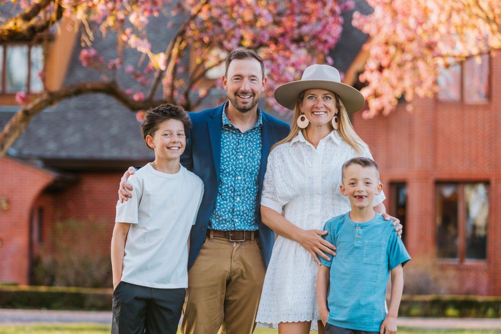 Happy family of four posing outdoors near a red barn with pink cherry blossom trees in spring.