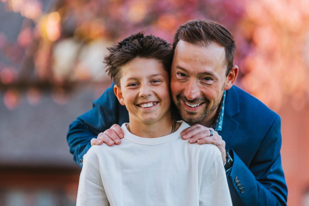A smiling man in a blue blazer hugs a happy young boy in a white shirt outdoors with fall foliage.
