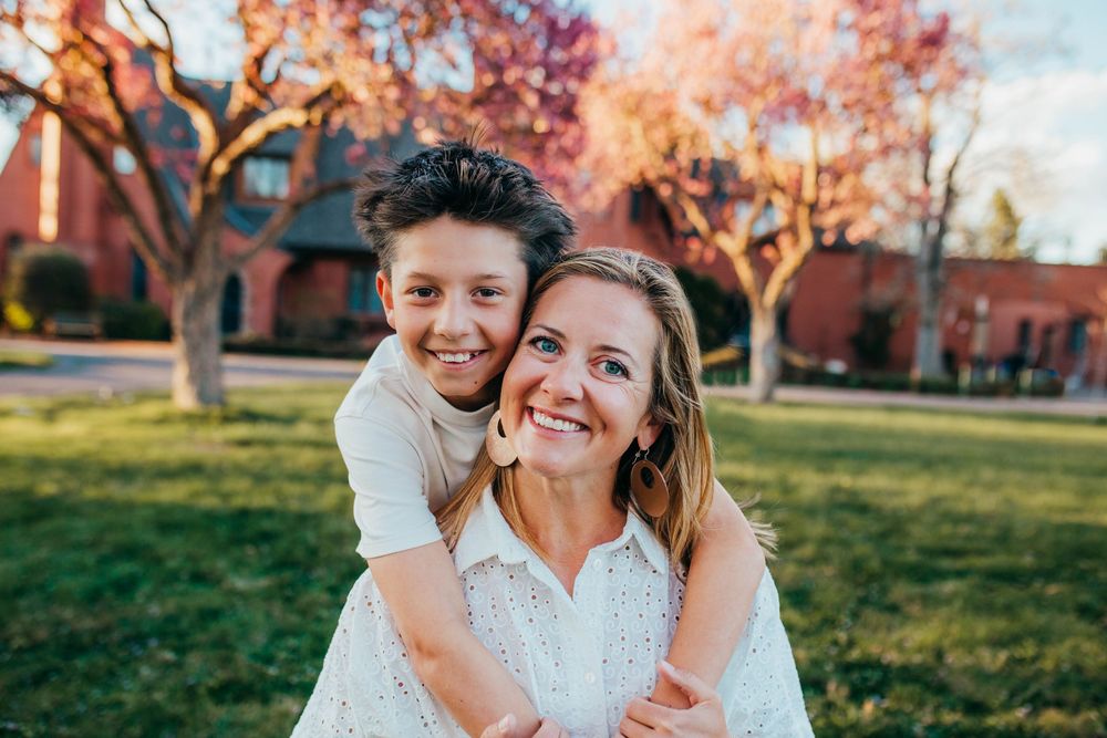 A smiling woman and young boy pose together outdoors near autumn trees and a brick building on a sunny day.