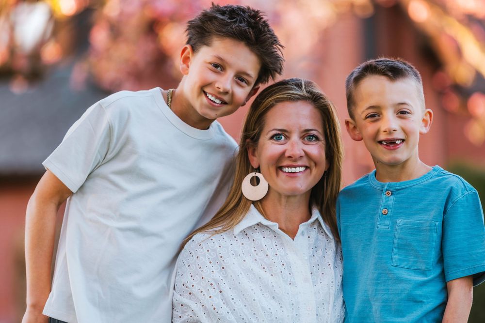 A smiling woman poses outdoors with two young boys, framed by pink blossoms in a warm family portrait.