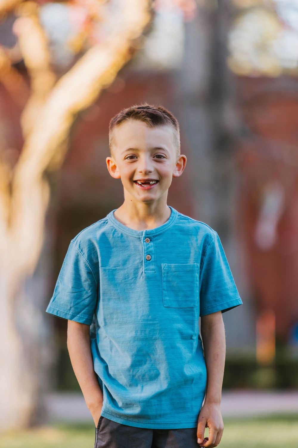 Smiling young boy with missing front teeth wearing a blue henley t-shirt standing outdoors in warm sunlight.