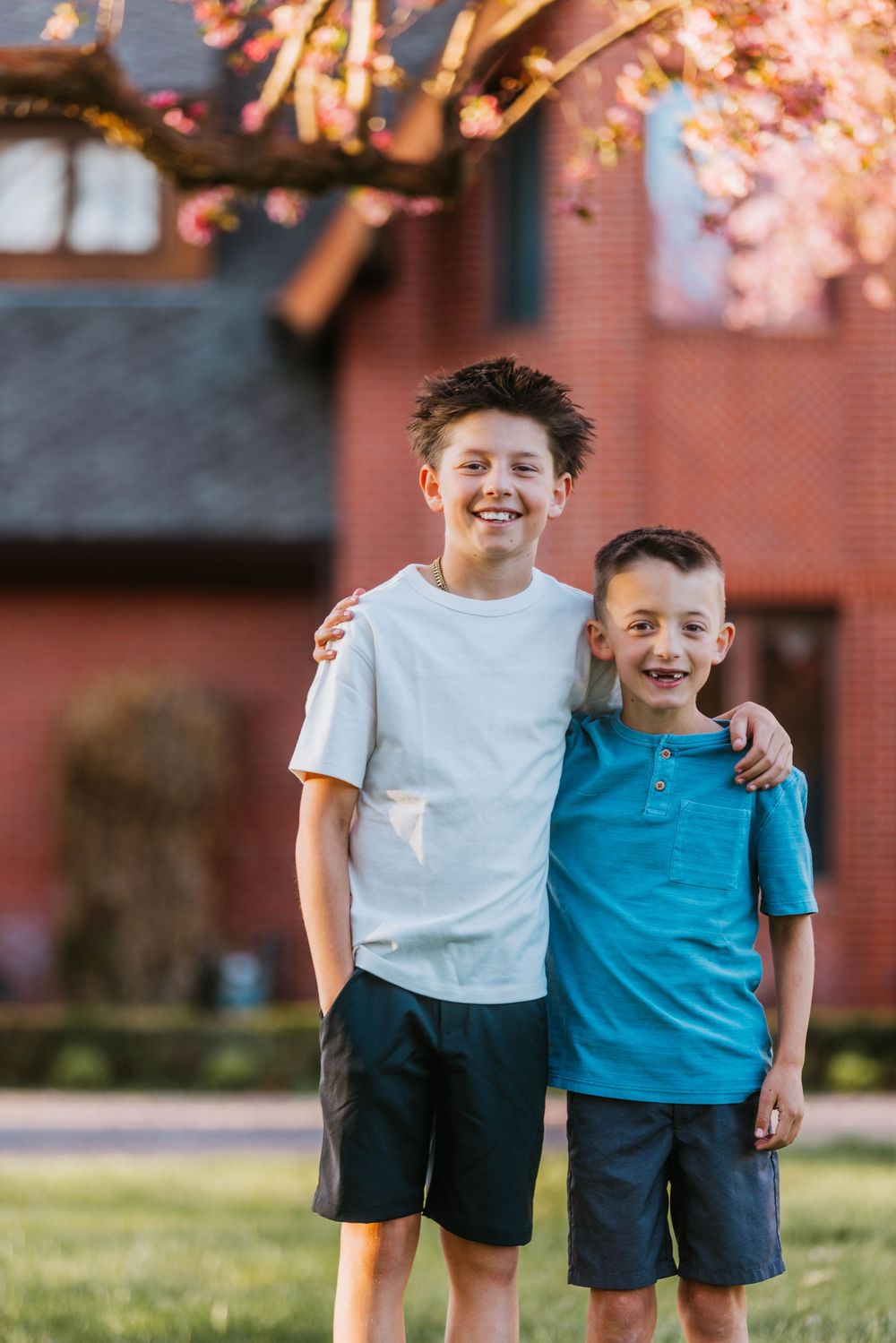 Two smiling boys in casual summer clothes embrace outdoors in front of a red brick house with blooming spring tree.