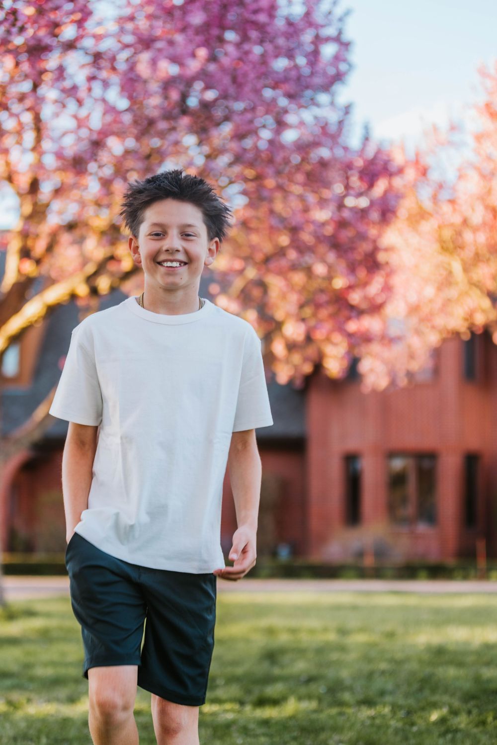 Smiling teenage boy in white t-shirt and dark shorts standing outdoors with pink flowering trees in background.
