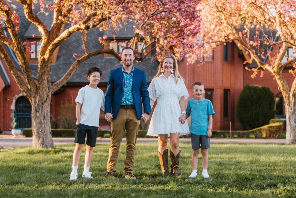 Family of four holding hands outdoors near pink flowering trees and a red barn at golden hour.