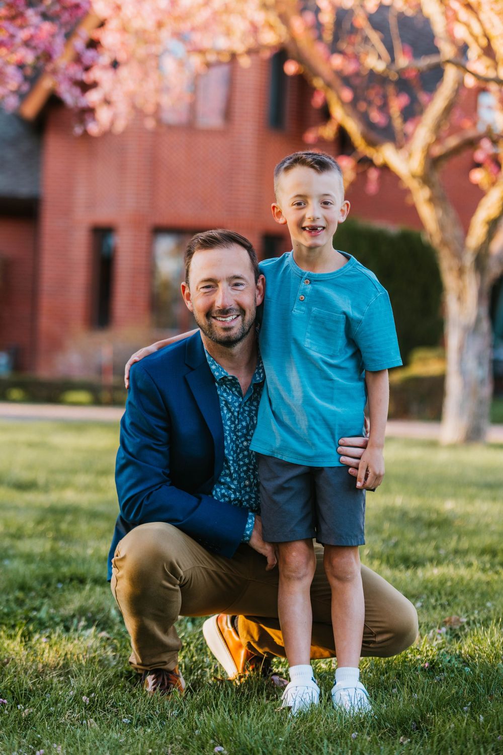 A smiling man in a blue blazer crouches beside a happy young boy in a teal shirt on a green lawn.