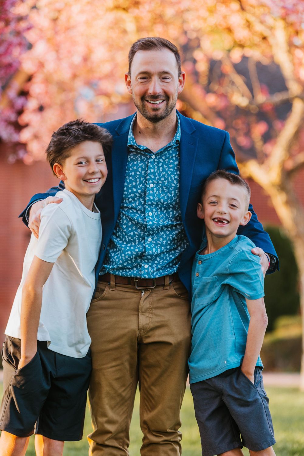 Smiling man in blue jacket hugs two young boys outdoors near blooming pink tree in warm sunlight.