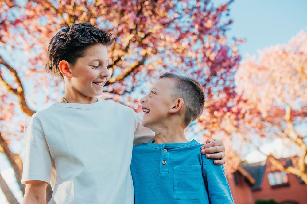 Two smiling boys share a joyful moment outdoors under a blooming pink tree in warm sunlight.