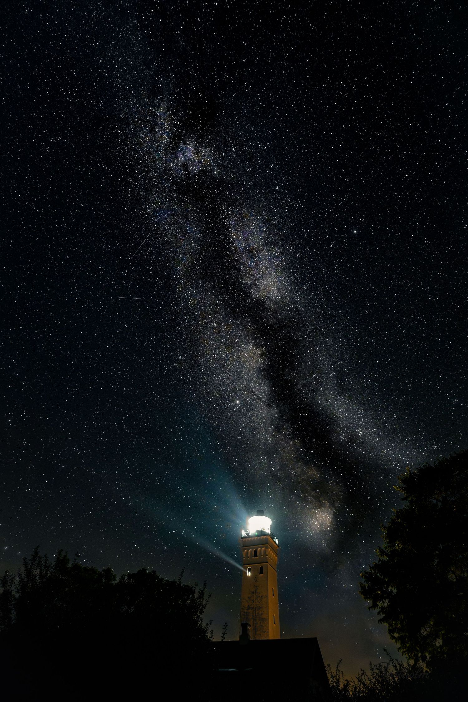 Illuminated lighthouse beam shining upward toward the Milky Way galaxy on a clear starry night sky.