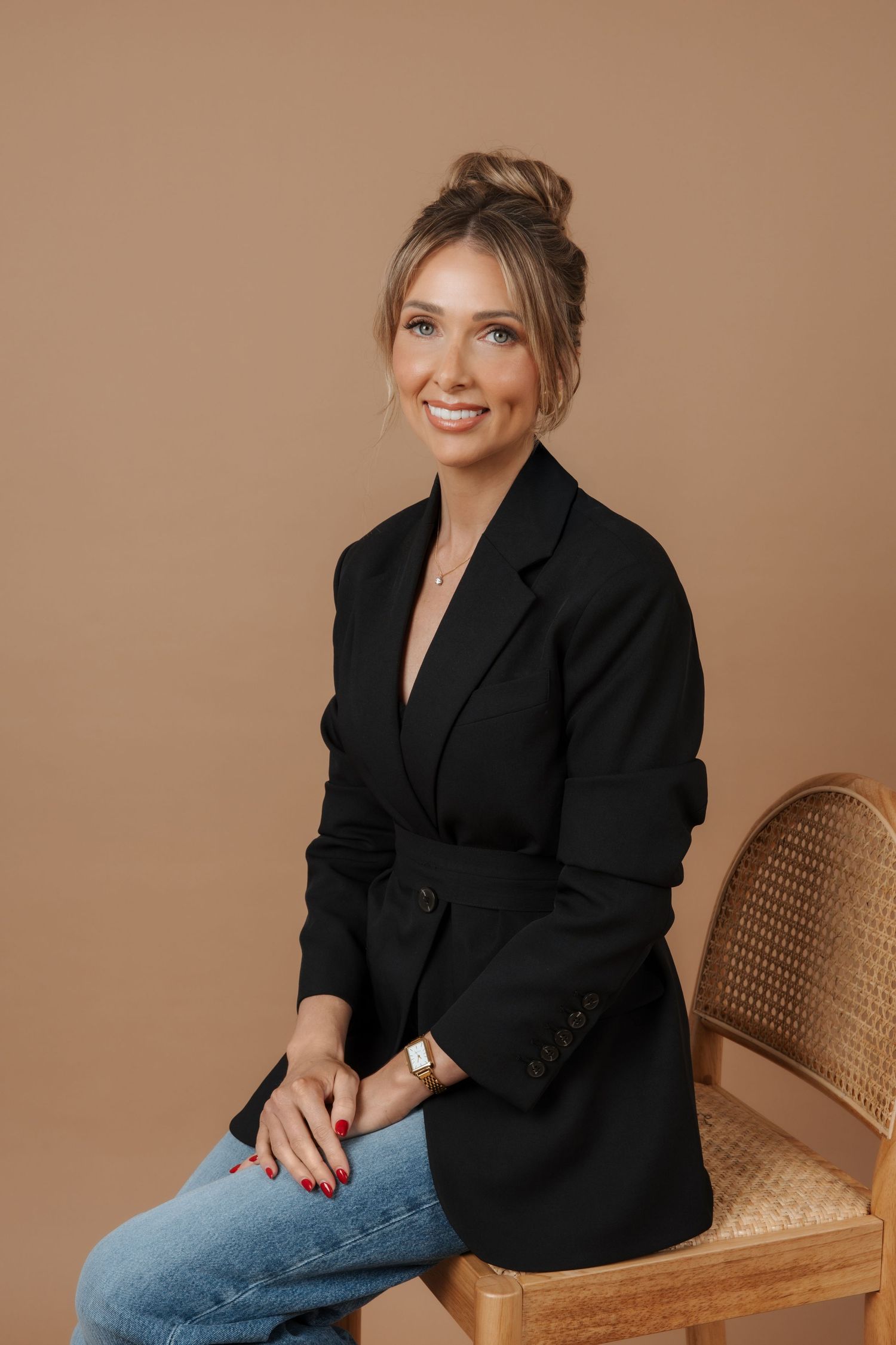 Smiling woman in black blazer and jeans sitting on a wicker chair against a warm beige background.
