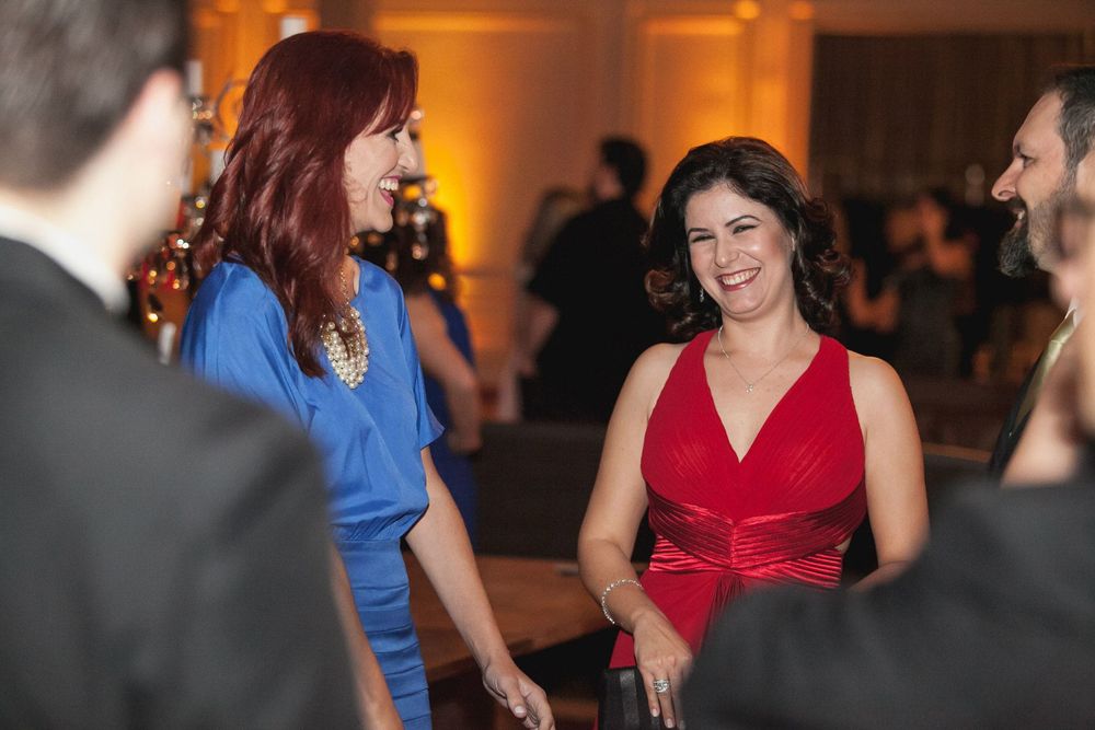 Two women in elegant blue and red dresses smiling and conversing at a formal evening event.