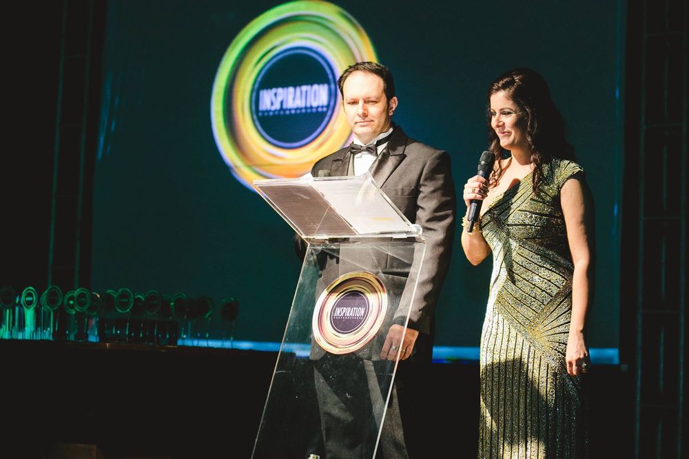 A man and woman hosting an awards ceremony on stage, with a podium and colorful logo backdrop.