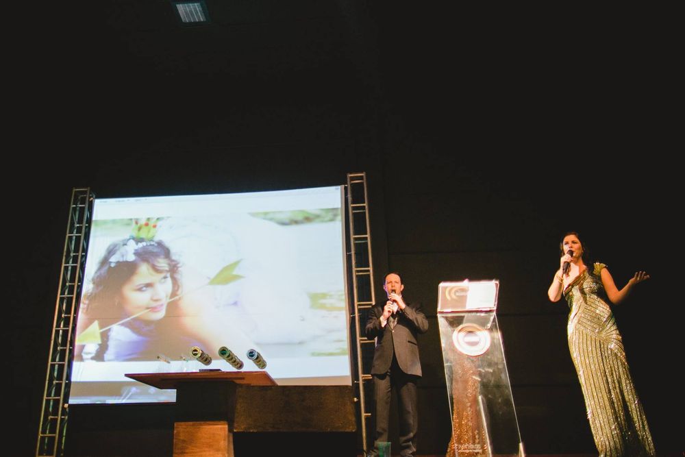 Two presenters on stage with microphone at award ceremony, large screen displaying portrait photo in dark auditorium.