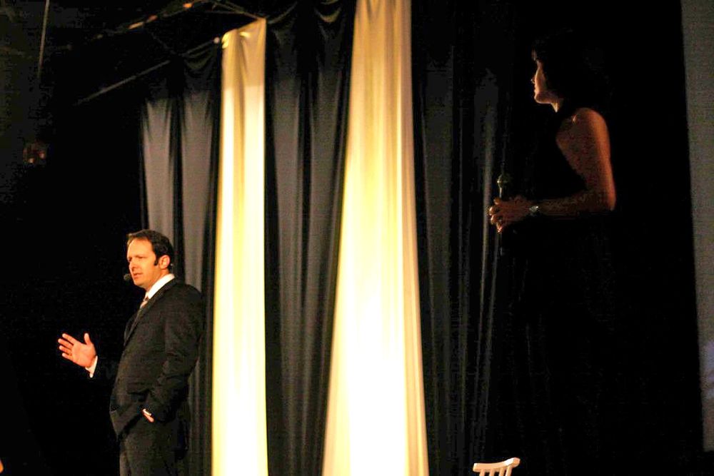 Two performers on a dimly lit stage with black and white curtains during a theatrical performance.