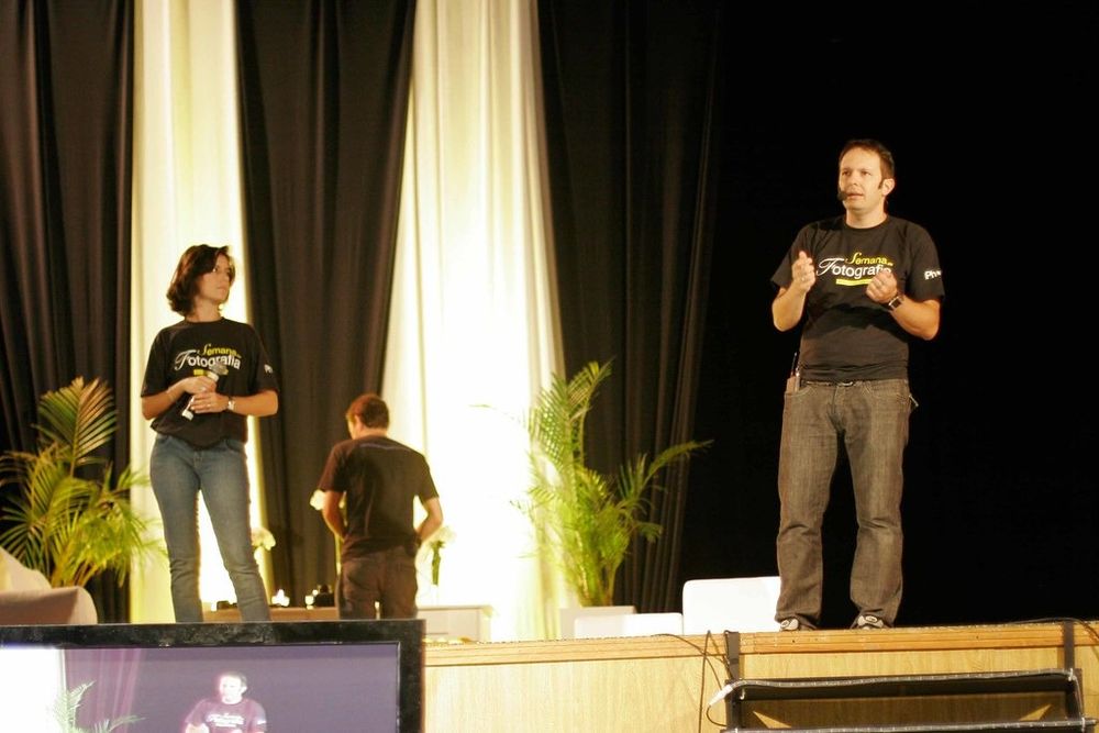 Three people on a stage with green plants, two wearing matching black t-shirts at an indoor event or conference.