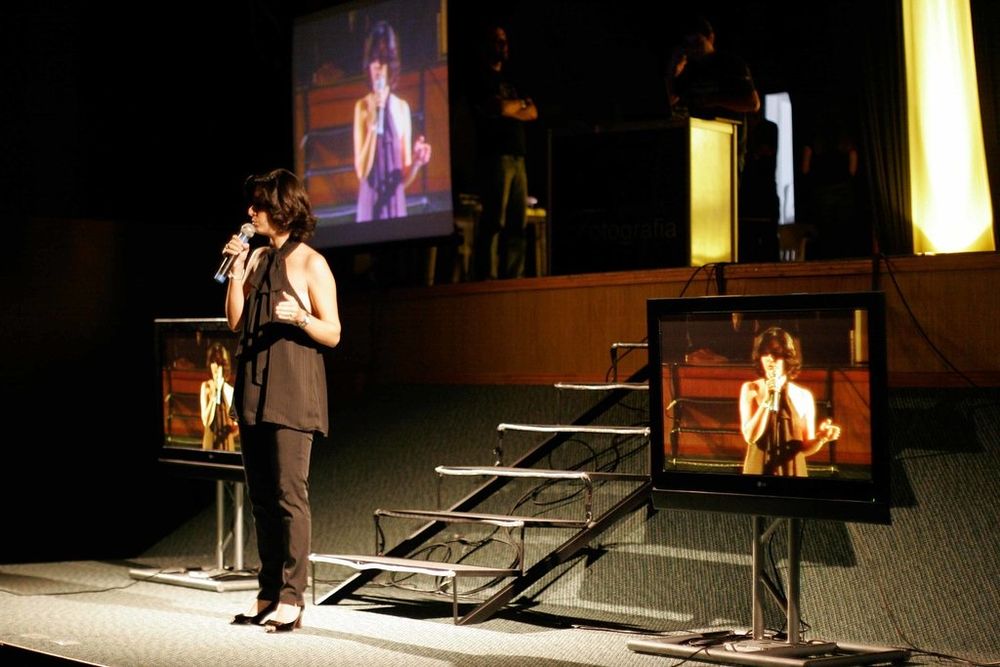 Performer speaks on stage surrounded by multiple TV monitors displaying live feed at a theatrical event.