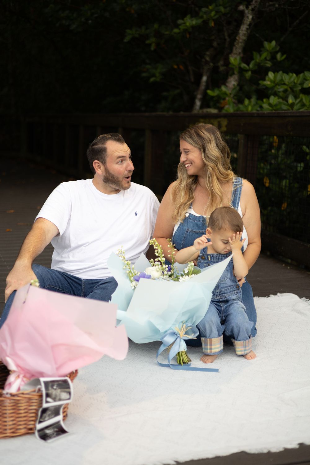 A couple sits on a white blanket outdoors with their baby, surrounded by blue and pink tissue paper and ultrasound photos.