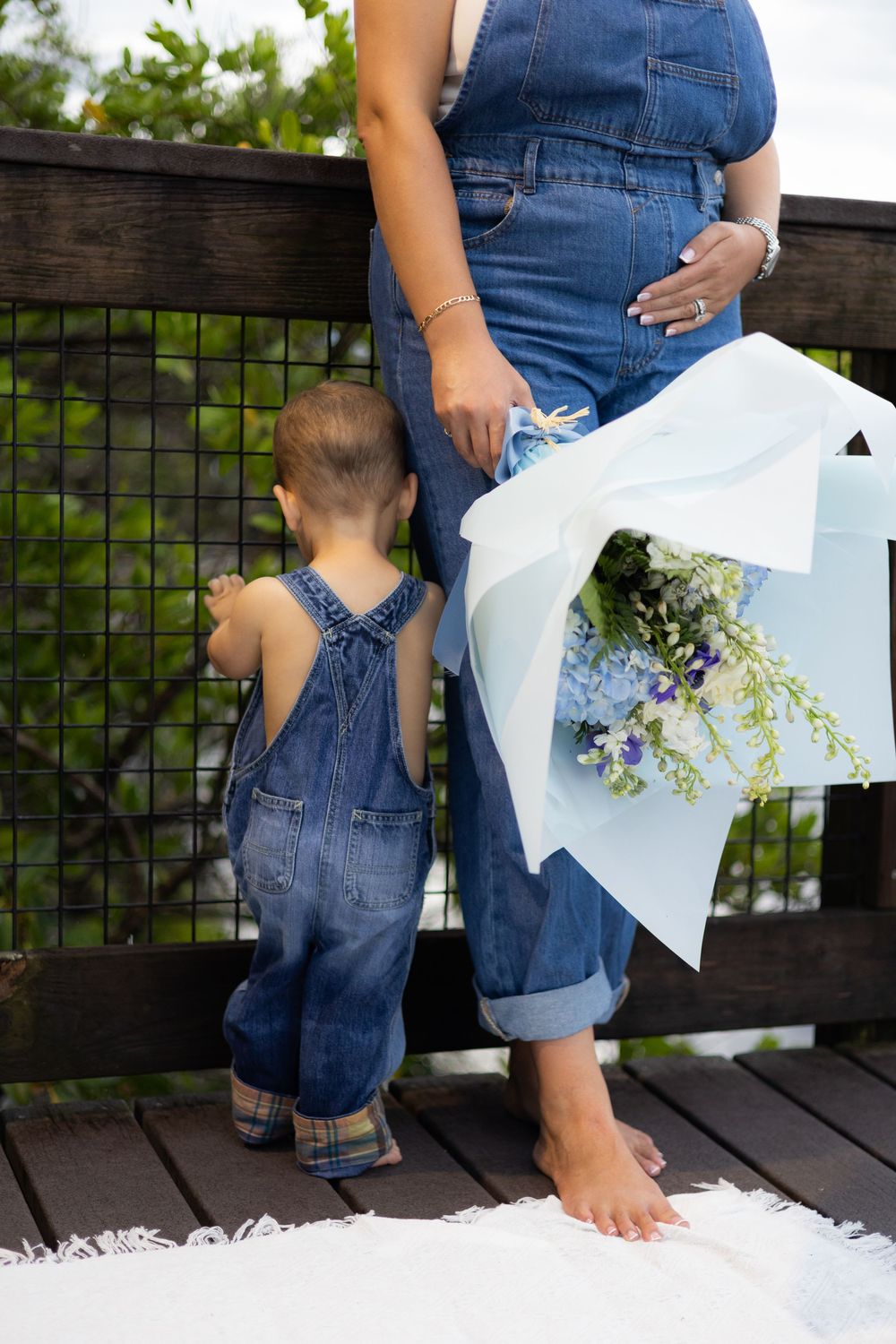 Pregnant woman in denim overalls holds flower bouquet beside toddler in matching denim overalls on wooden deck.