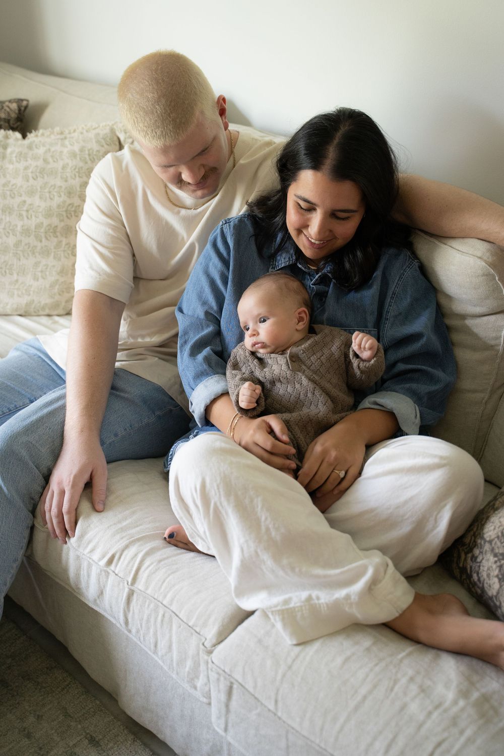 A young family sits on a couch, smiling at their newborn baby dressed in a brown outfit.