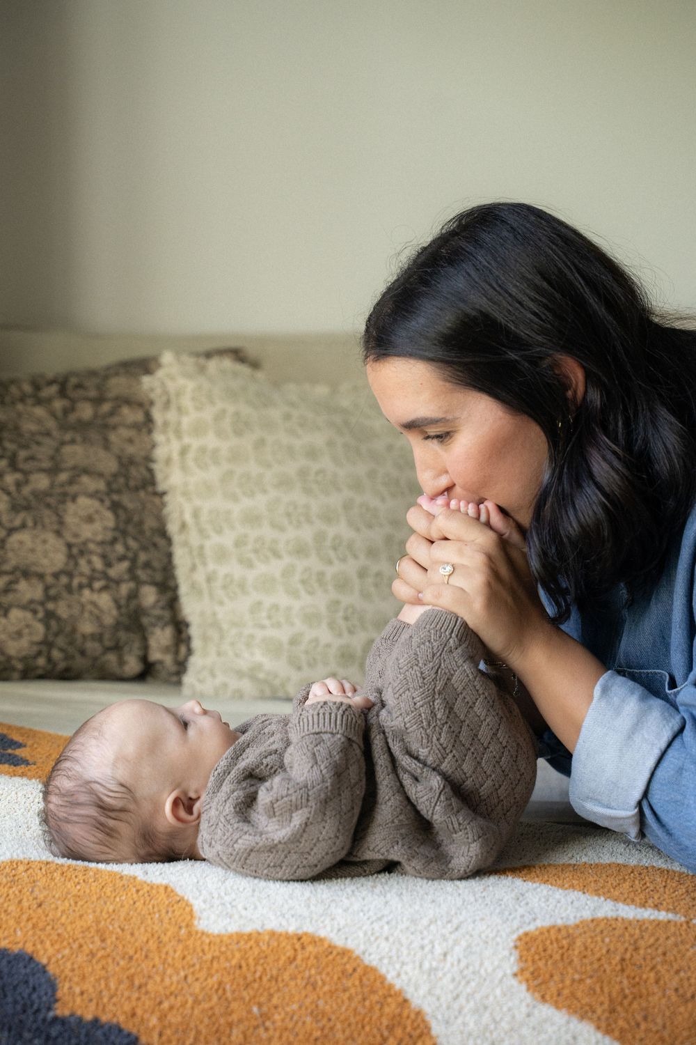 A loving mother kisses her baby's hands while the infant lies on a colorful blanket on a cozy couch.