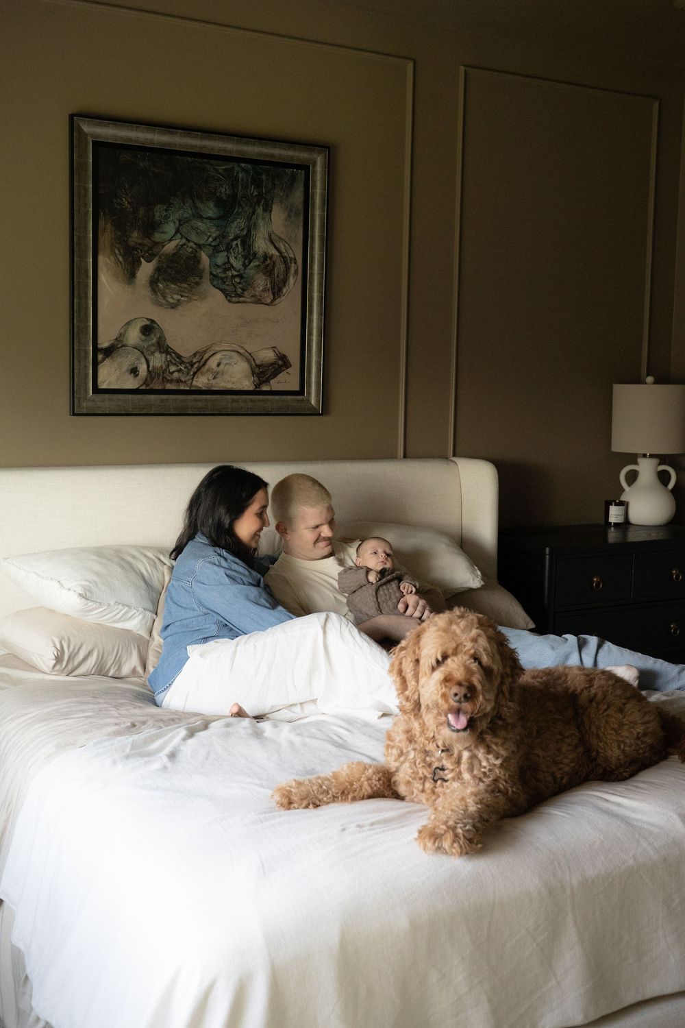 A couple relaxes on a white bed with their newborn baby and fluffy golden doodle dog in a cozy bedroom.