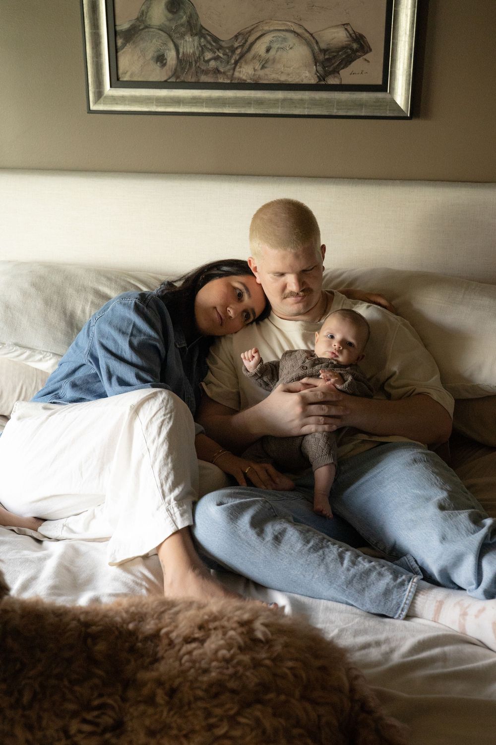 Young family with a blonde father and dark-haired mother cuddling their newborn baby on a bed.