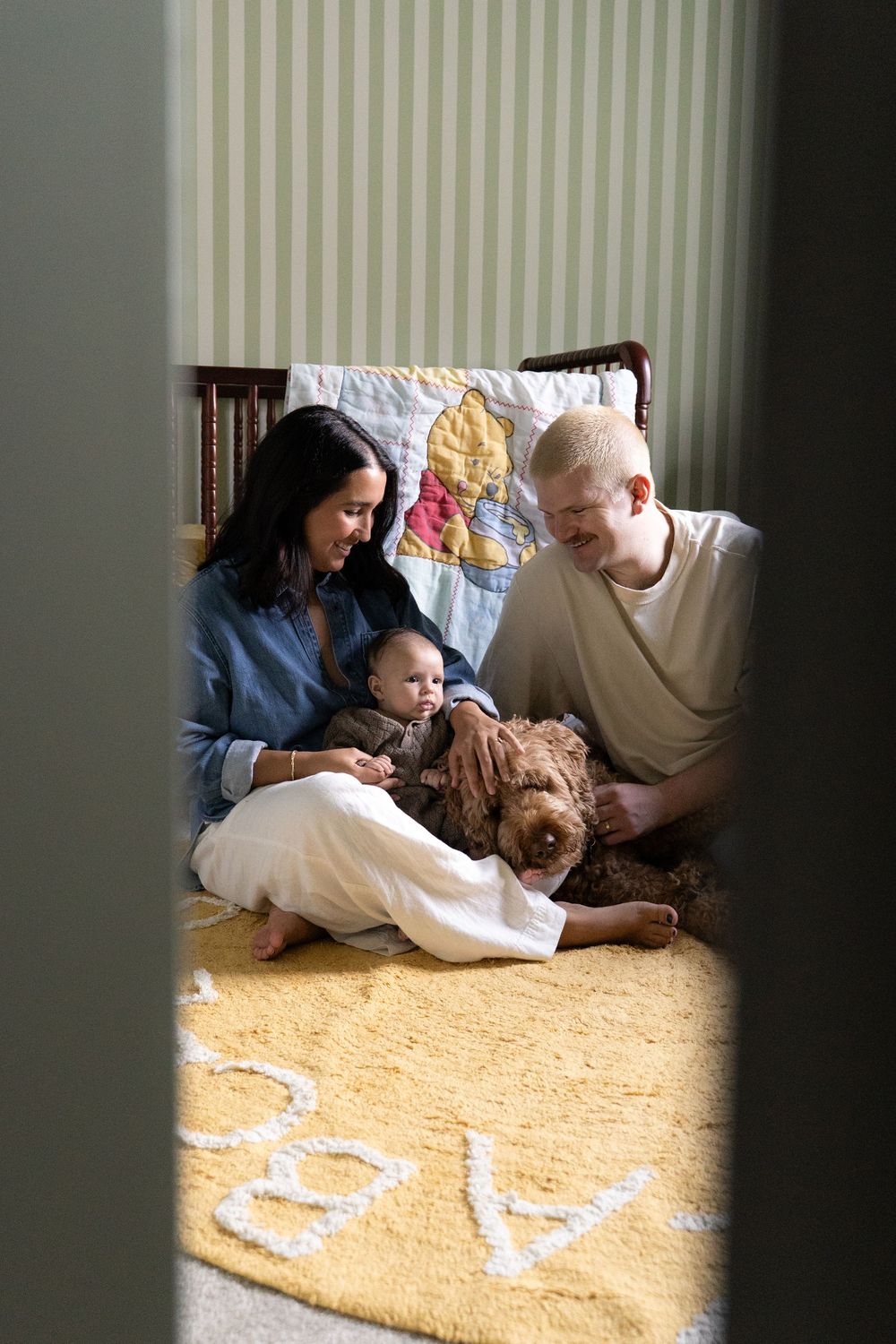 A couple sits on a nursery floor with their baby and a fluffy brown dog on a yellow ABC rug.