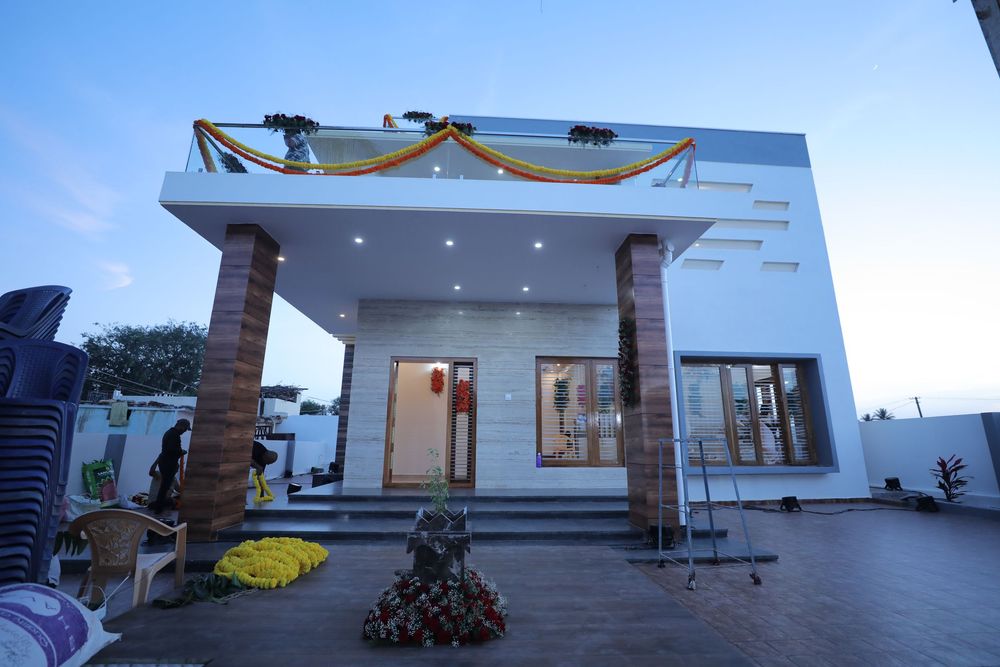 Modern white single-story house with decorative marigold garlands and stone pillars at dusk, ready for housewarming ceremony.