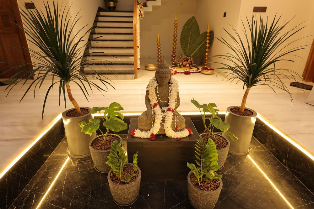 Indoor decorative display featuring a Buddha statue adorned with flowers, surrounded by tropical potted plants and warm LED lighting.