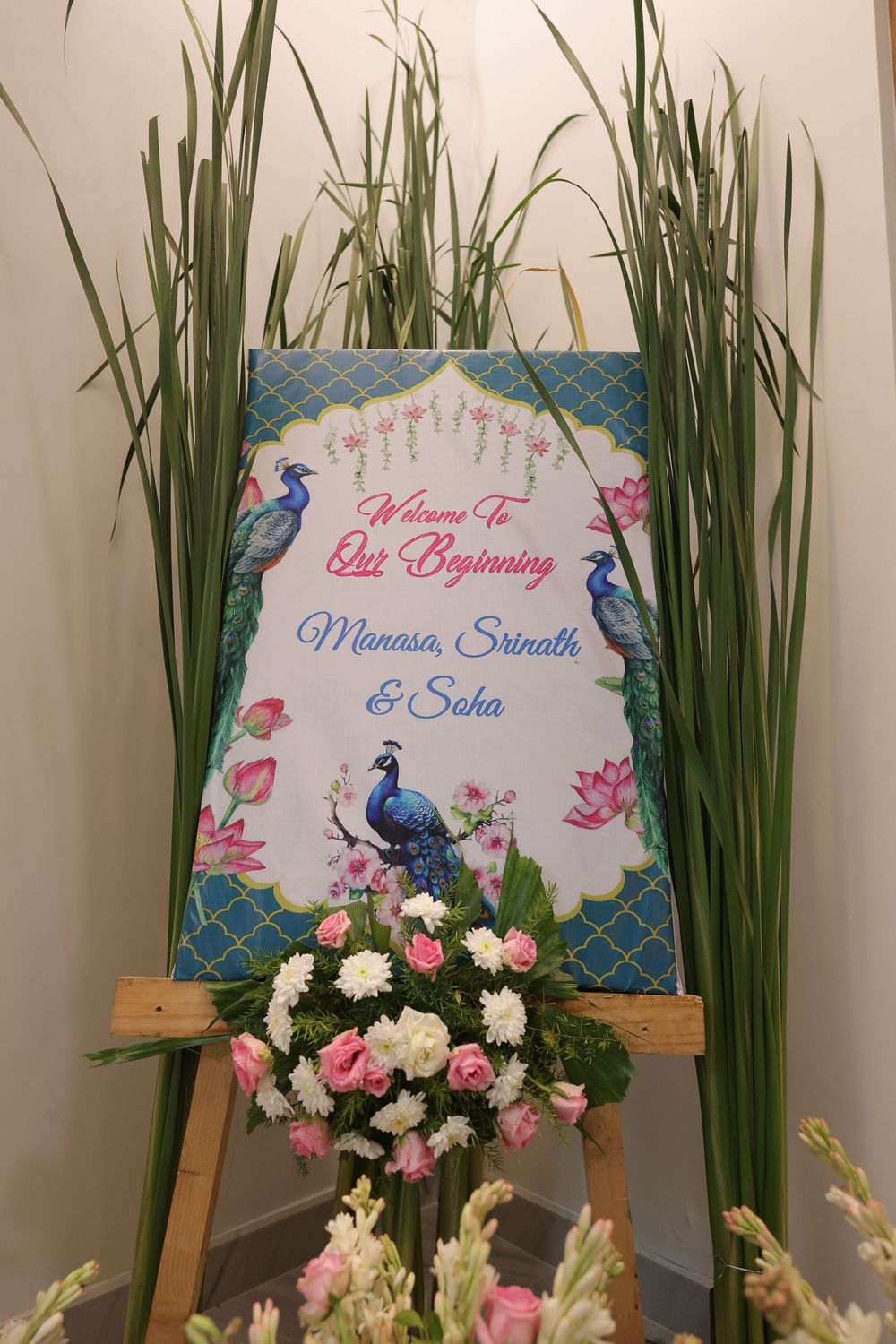 Elegant wedding welcome sign for Manasa, Srinath & Soha on wooden easel with peacock design, pink roses and white flowers.