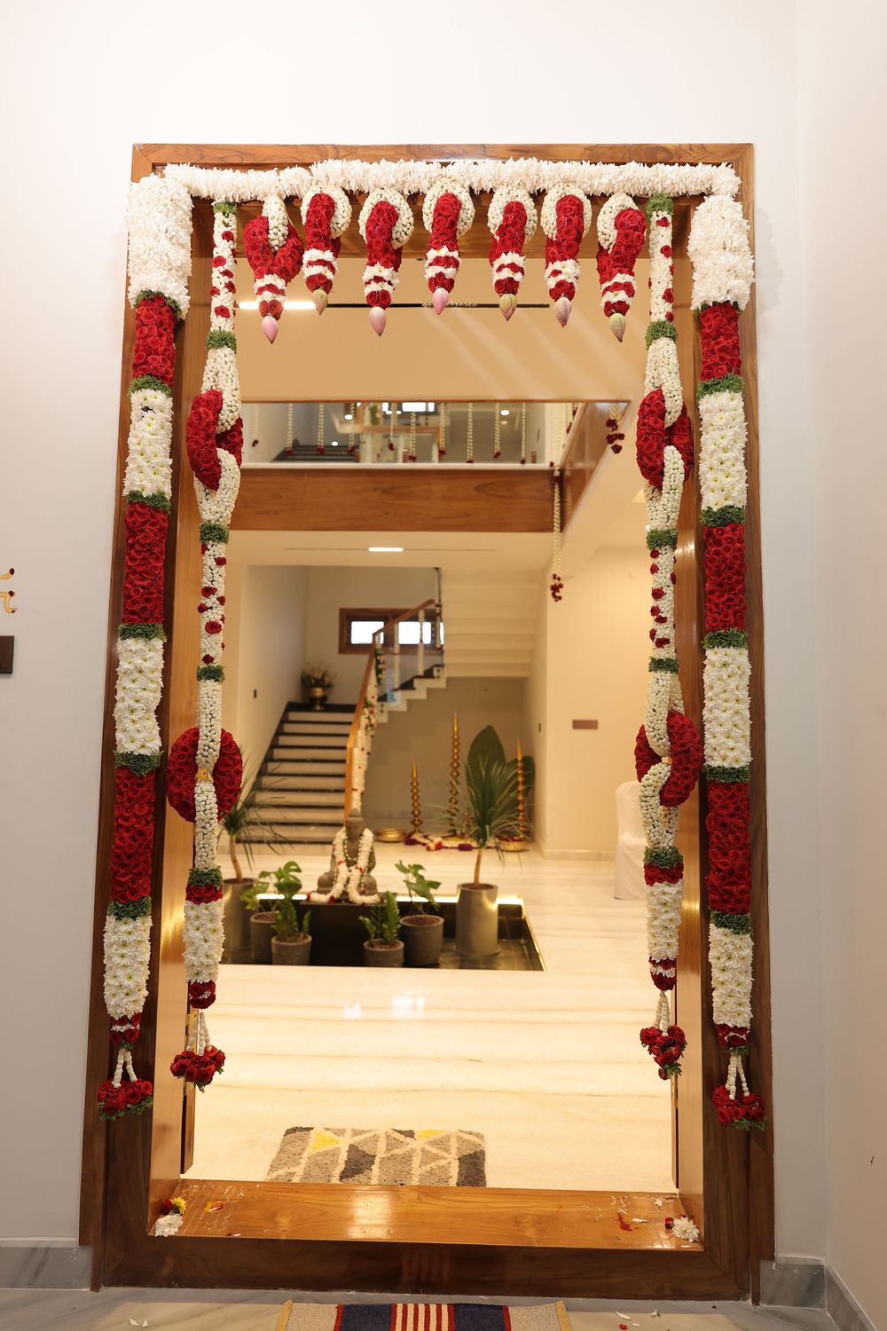A wooden doorframe adorned with vibrant red and white flower garlands leading into a modern Indian home interior.