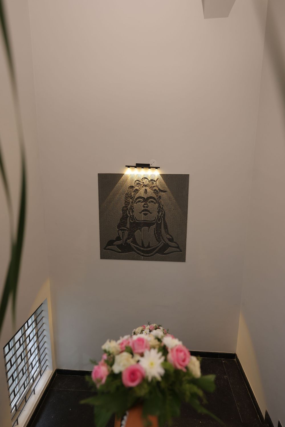 Wall-mounted Adiyogi Shiva relief sculpture with spotlight above fresh pink and white flower arrangement below.