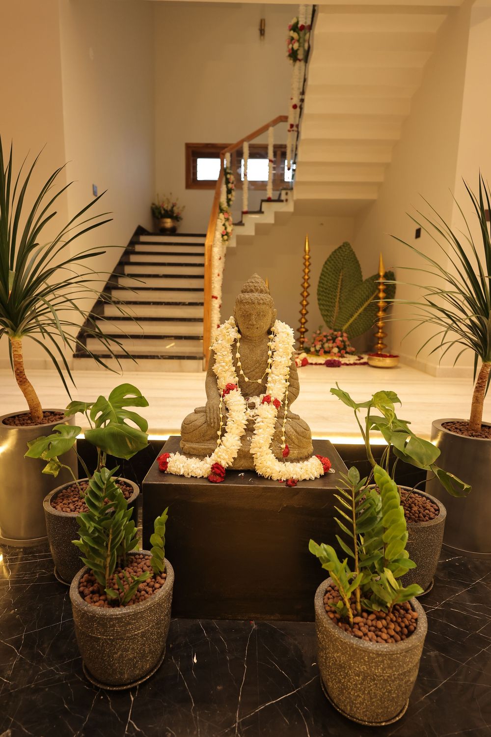 Buddha statue adorned with floral garlands surrounded by tropical potted plants in an elegant indoor foyer with staircase.