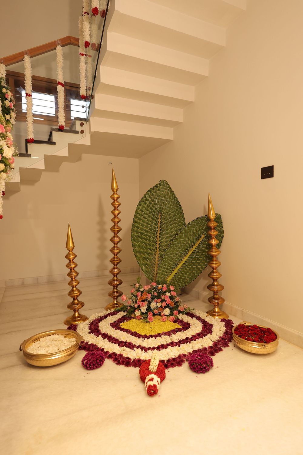 Traditional South Indian home decor with brass lamps, floral rangoli, betel leaf, and flower arrangements near staircase.