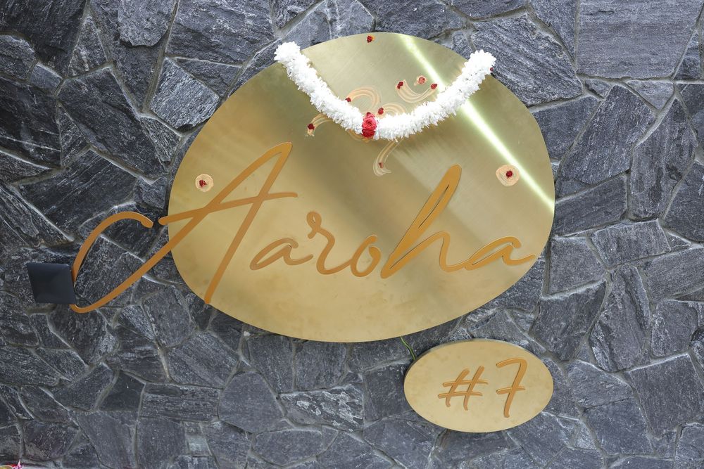 Gold oval nameplate reading Aaroha with number 7 plaque mounted on gray stone wall with floral garland.