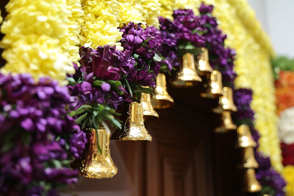 Gold bells and purple flower bouquets hang from a colorful floral arch with yellow and purple garlands.
