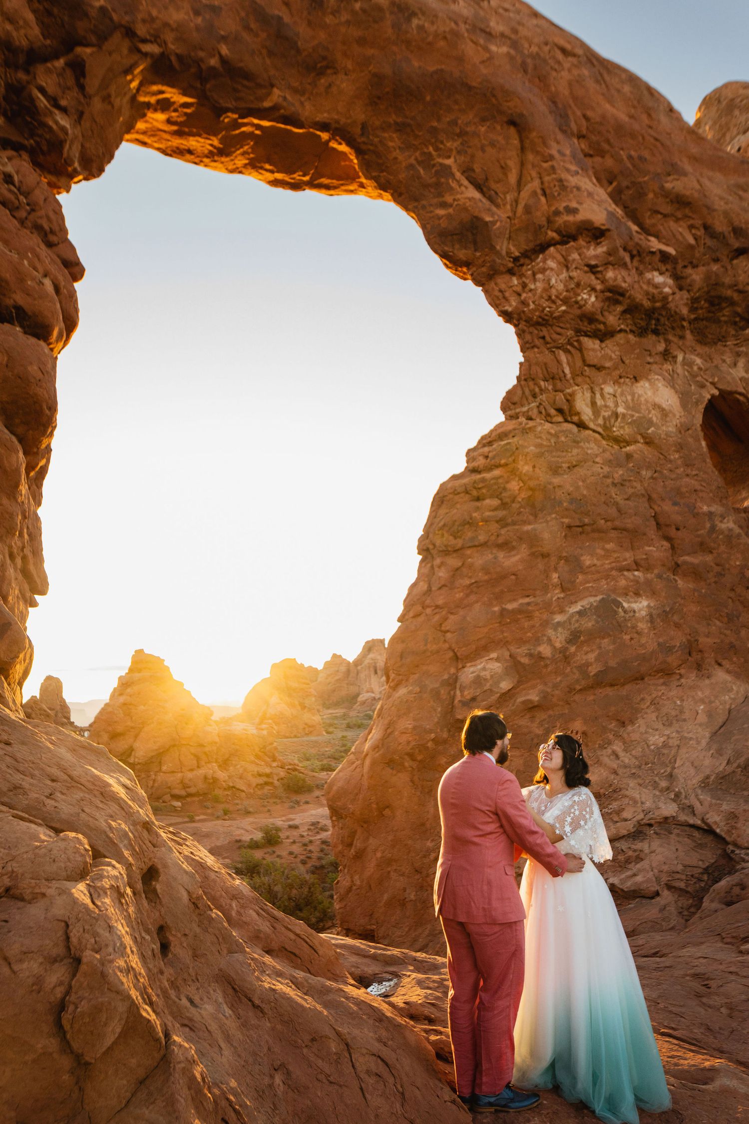 Couple embracing under a natural sandstone arch at sunset in the desert.