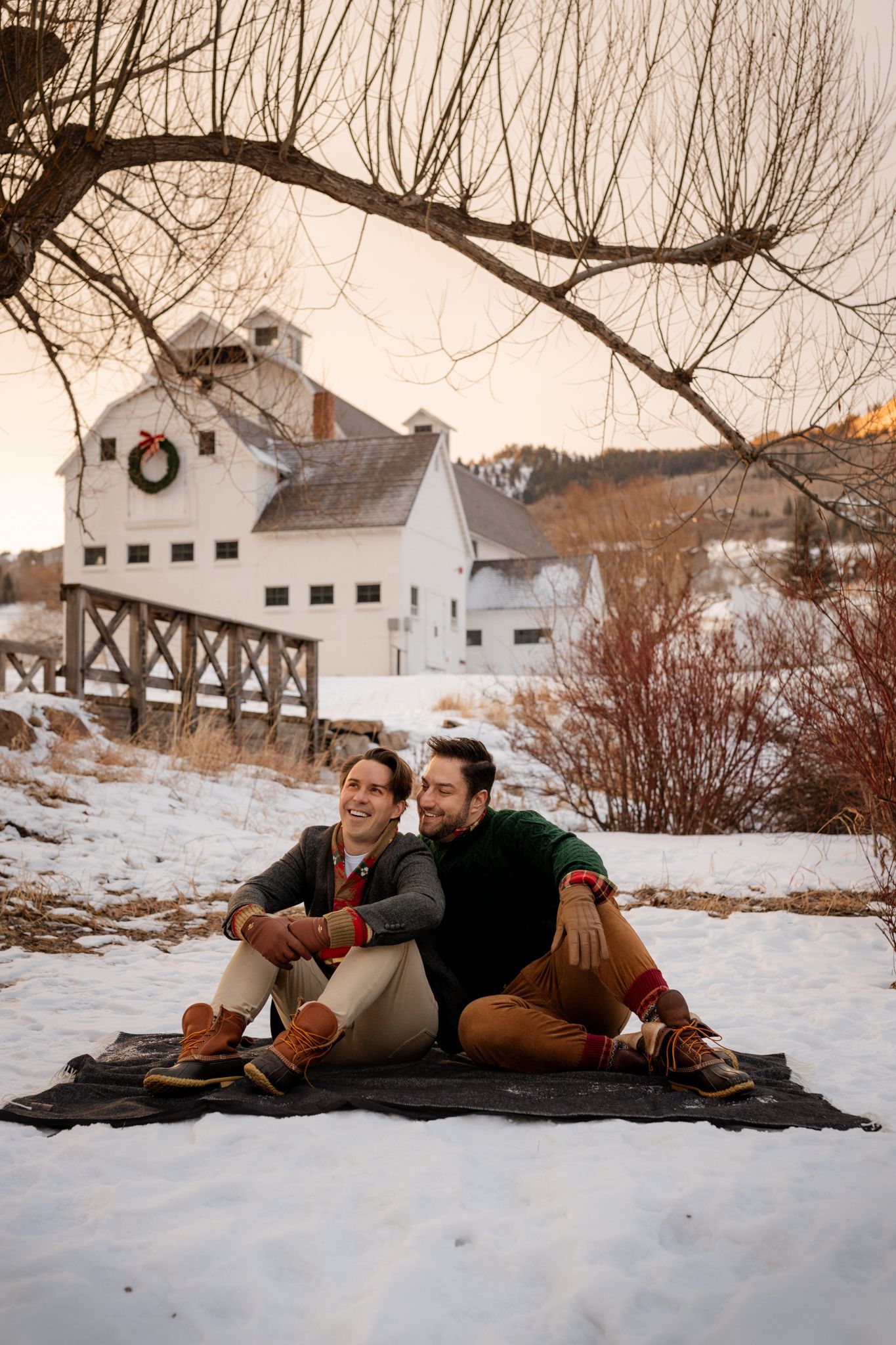 Two men sitting on a blanket in the snow, smiling together with a white barn decorated for Christmas behind them. LGBTQ wedding photography.