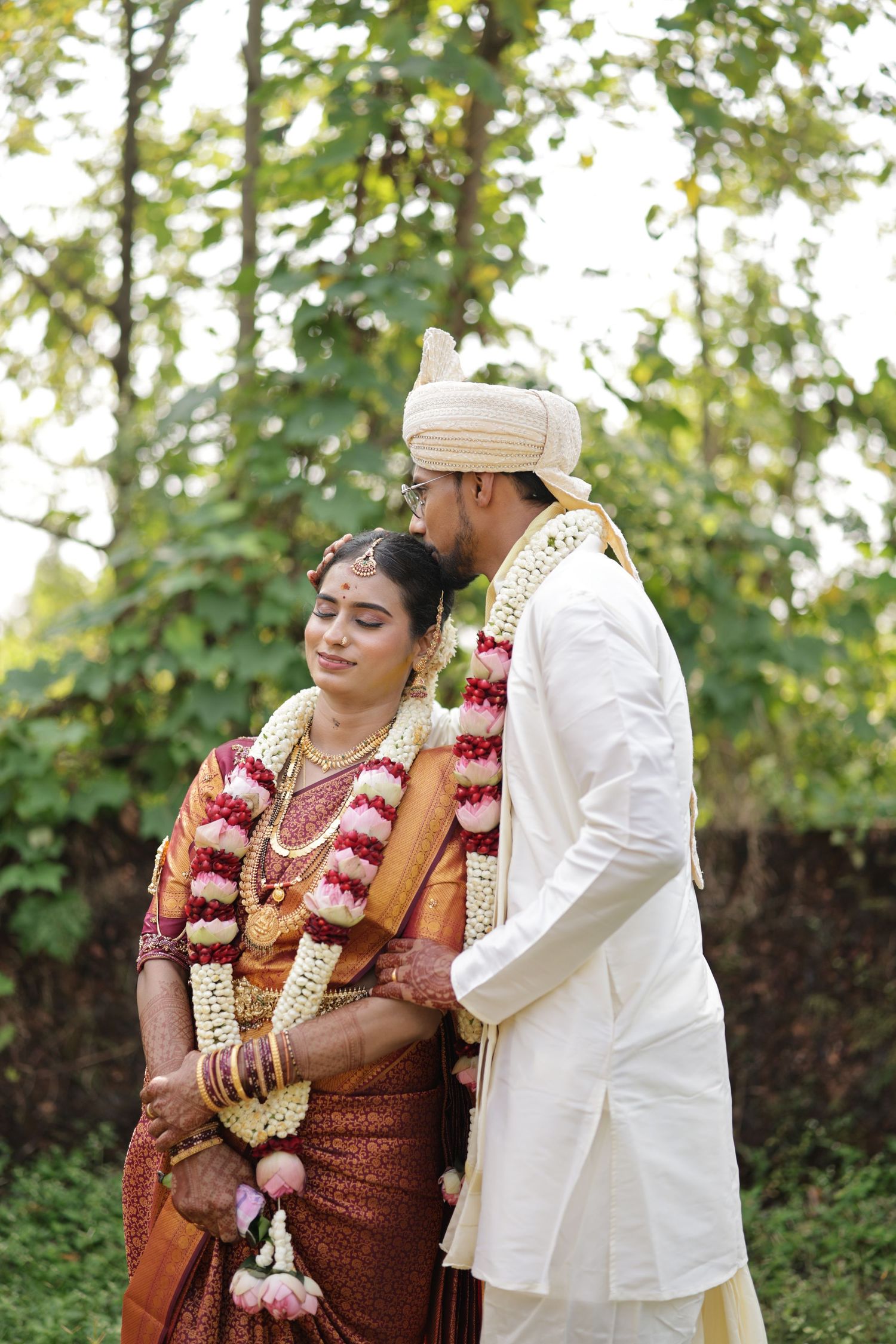 South Indian couple in traditional wedding attire sharing a joyful moment outdoors with lush greenery backdrop.