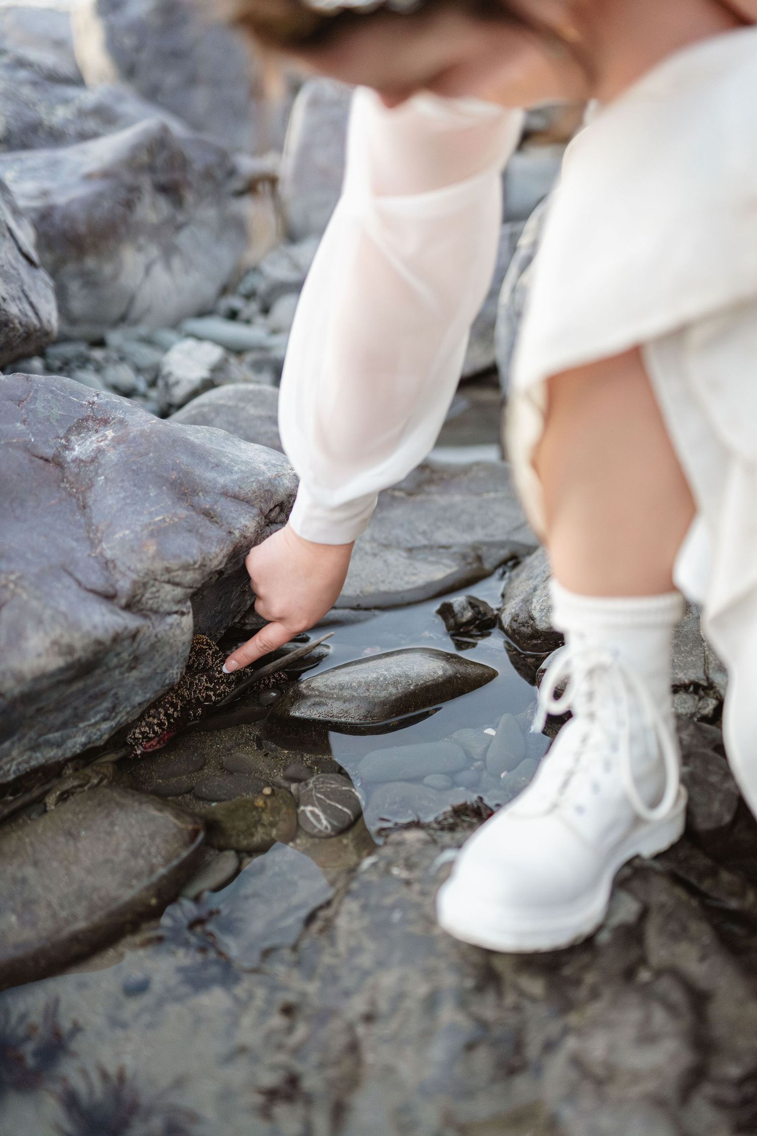 Bride in white dress and sneakers crouches to touch tide pool rocks at shoreline.