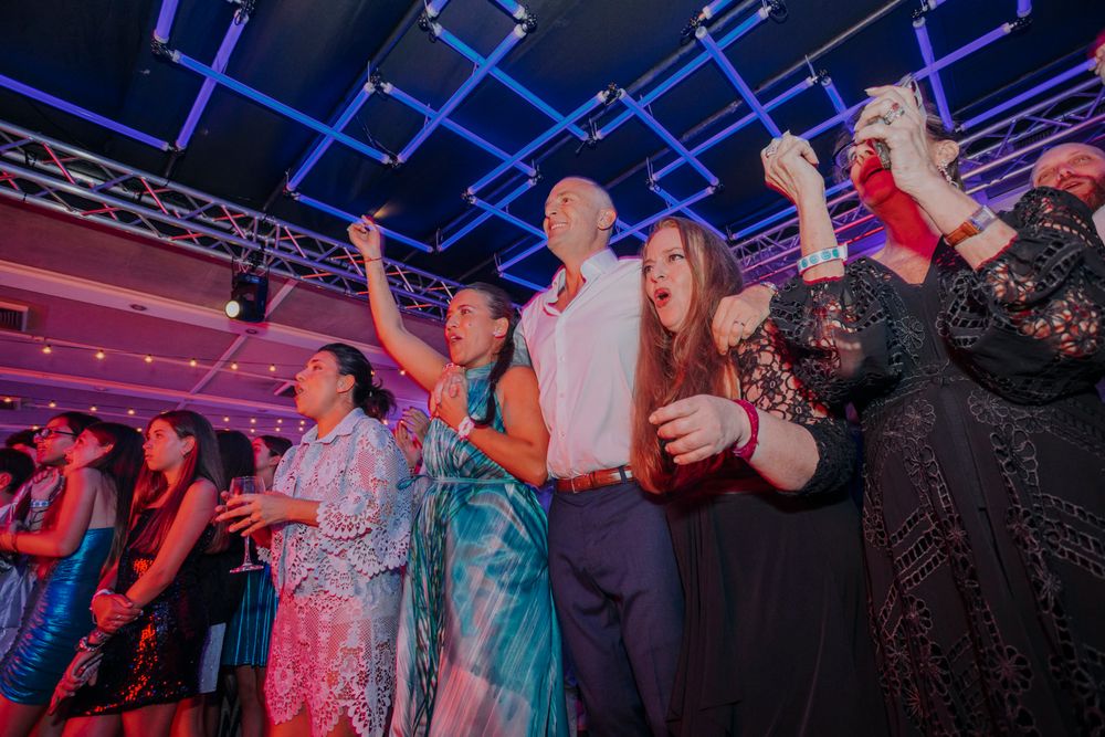 Excited crowd dancing and celebrating at a vibrant indoor party event with colorful pink and blue lighting.