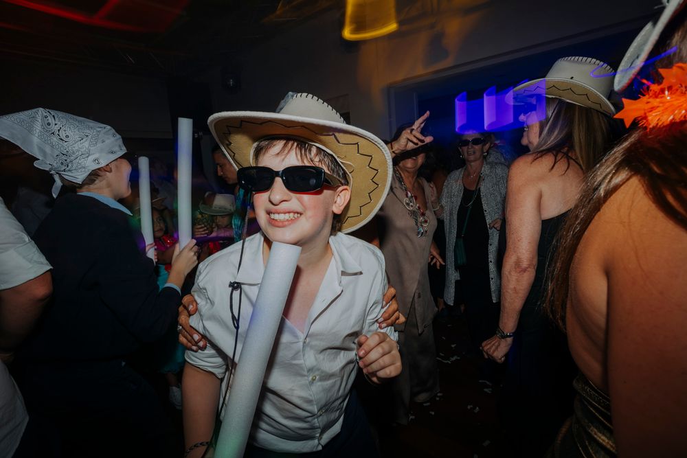Person in cowboy hat and sunglasses dancing at a party, holding glow sticks in a crowded nightclub.