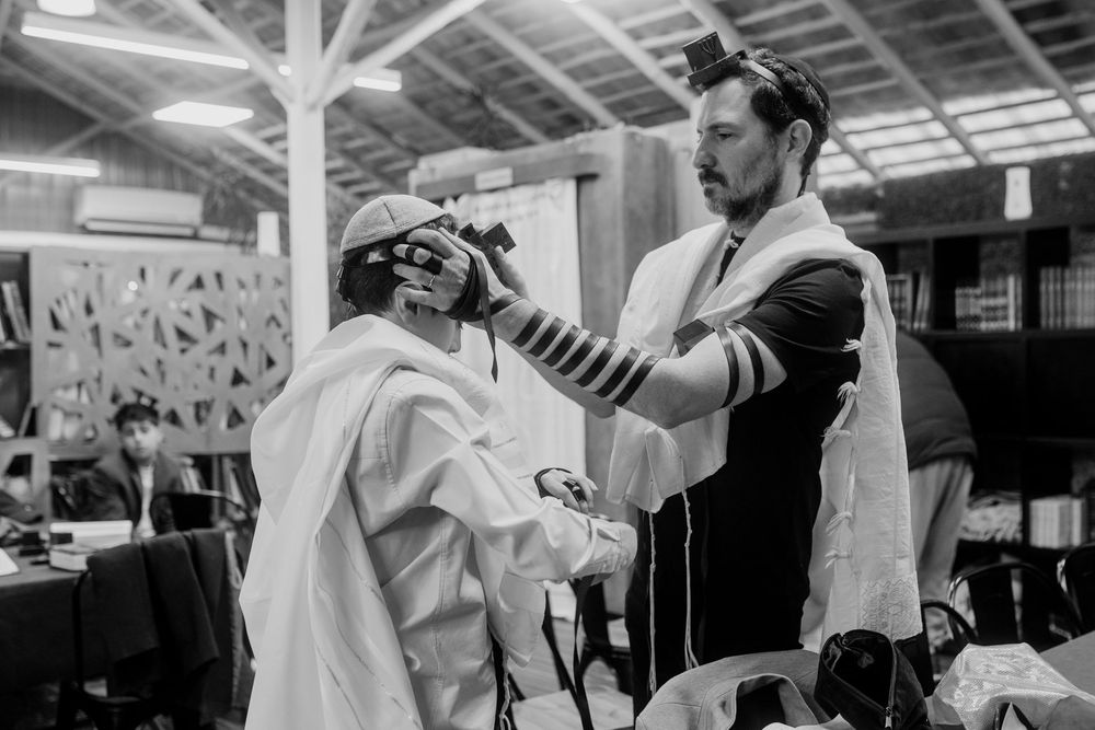 Jewish man in tallit helps another with tefillin wrapping during morning prayer service in synagogue.