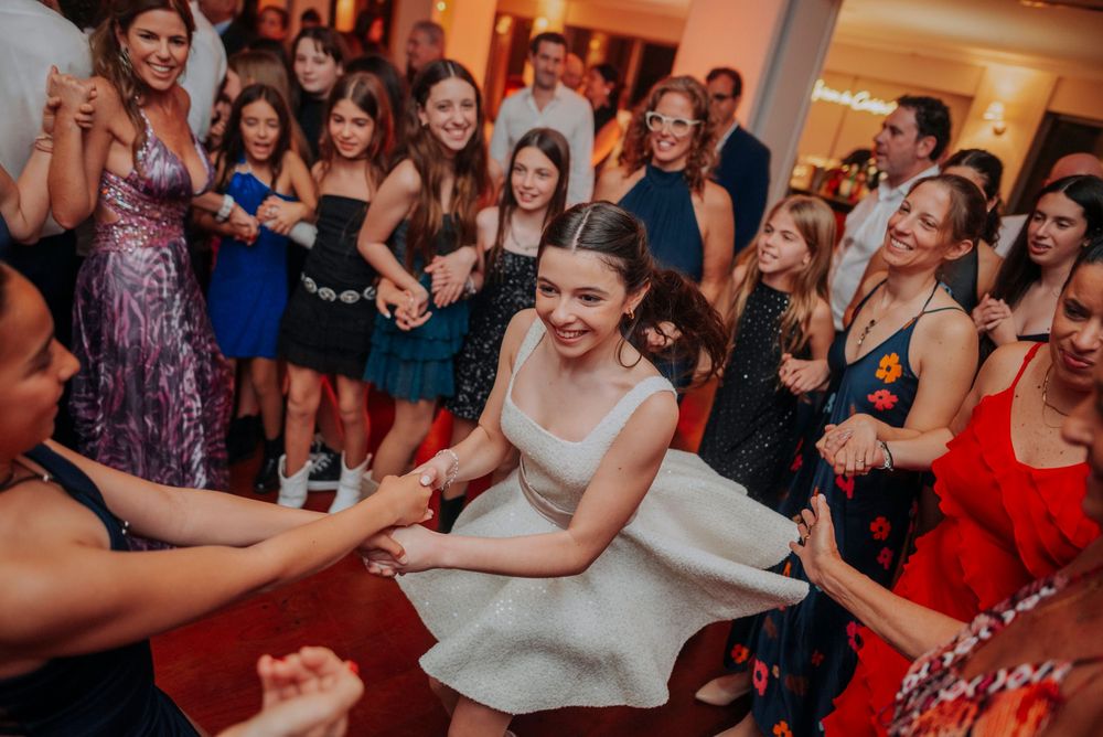 Young girl in white dress twirling joyfully on dance floor surrounded by smiling guests at a festive party.