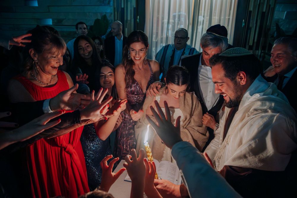 Group of people gathered around candles at a Jewish celebration, smiling and reaching toward the flame.