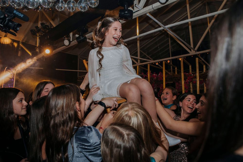 Young girl in white dress lifted by cheering friends at a festive Bat Mitzvah party celebration.