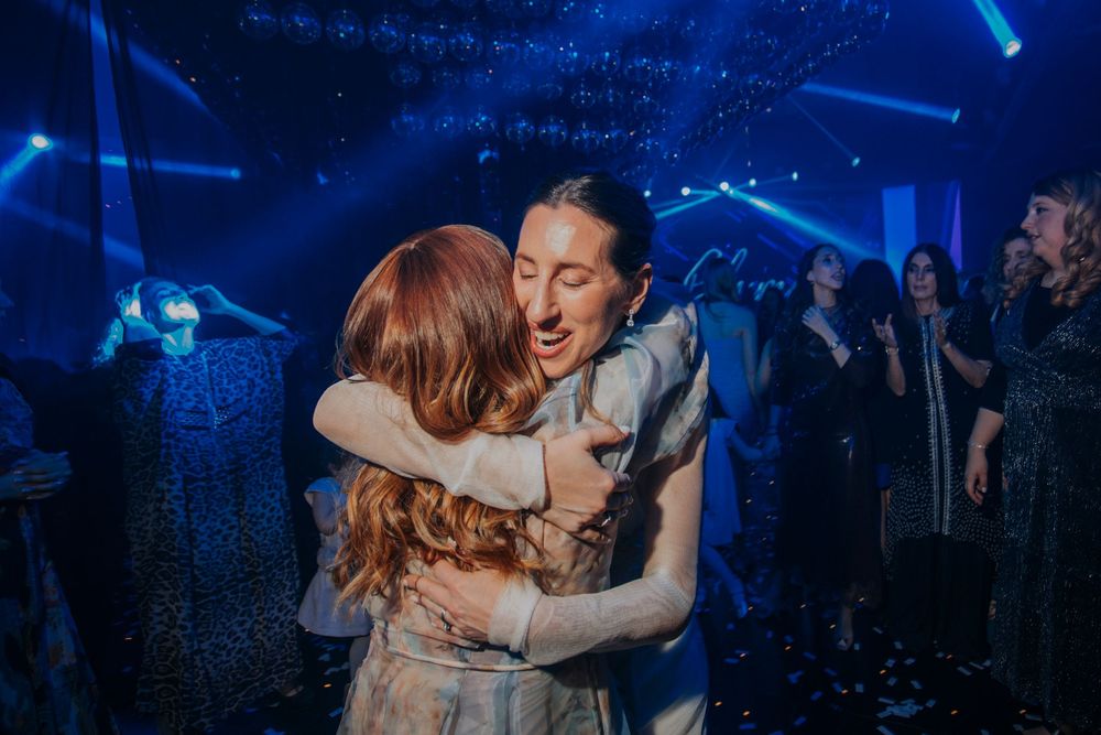 Two people share a warm hug on a dance floor with blue stage lighting and confetti at a celebration event.