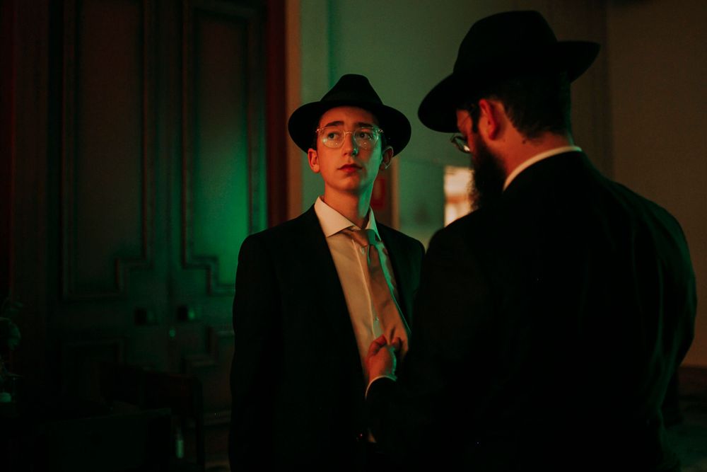 Two people in hats and formal wear having a conversation in moody green lighting.