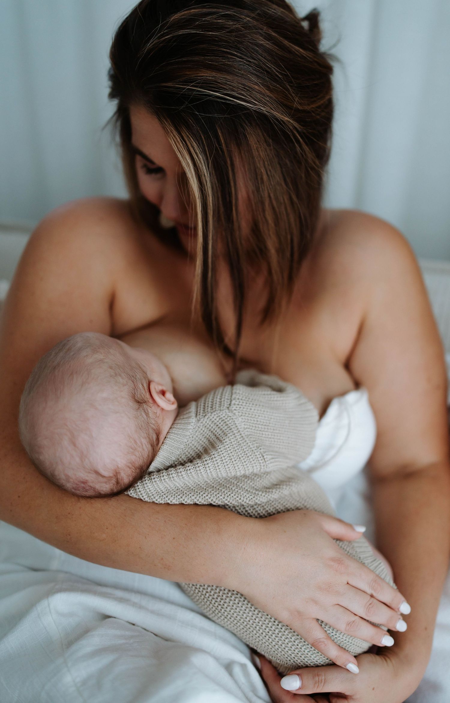A mother tenderly breastfeeding her newborn baby wrapped in a knit blanket while sitting on a white bed.