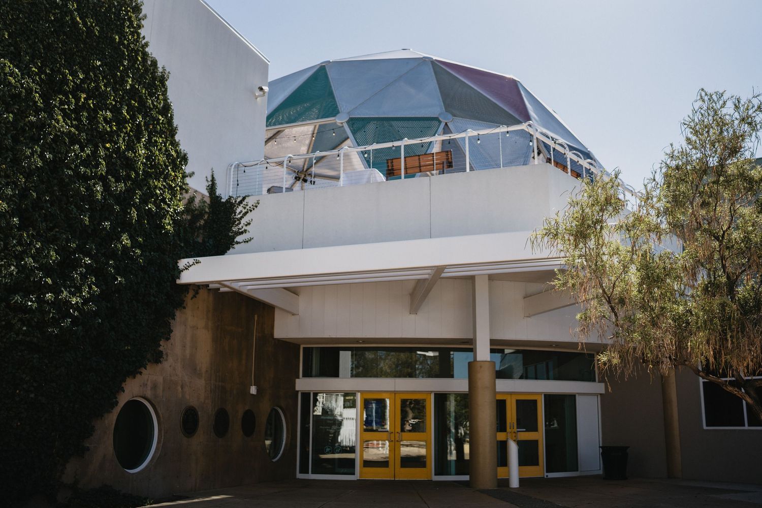 Modern building with geodesic dome roof, yellow-framed glass entrance doors, white facade, and circular windows on a sunny day.