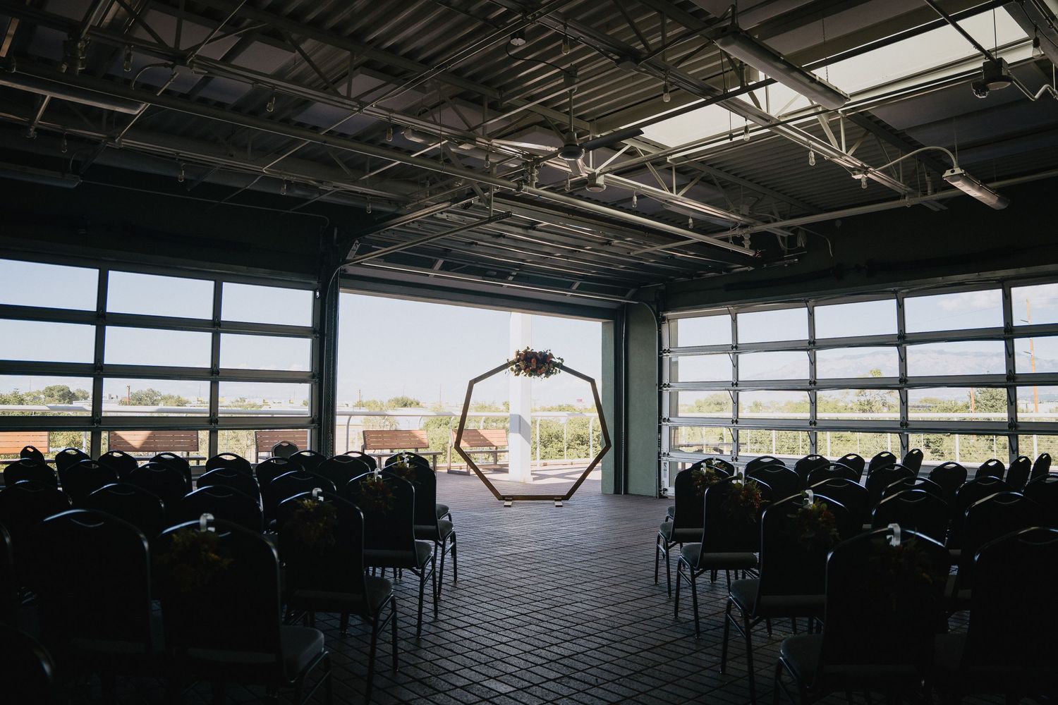 Industrial wedding venue with hexagonal arch, rows of chairs, and large garage doors opening to outdoor views.